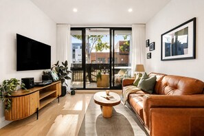 A modern living room with a flat-screen TV, a leather sofa, a coffee table, and a large window with a view of buildings. - The Vinyl Abode | Free Parking + 5-Minute CBD Walk (West Melbourne)
