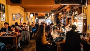 Een drukke bar met houten tafels en stoelen, gasten die zitten en staan, en planken vol flessen.