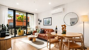 A modern living room with a leather sofa, wooden coffee table, and dining set. There is a large window with a view of buildings and greenery outside. - The Vinyl Abode | Free Parking + 5-Minute CBD Walk (West Melbourne)