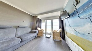 A modern living room with a grey sofa, wicker chairs, a small table, and a large window offering a view of the sea. - Stay in Middelkerke with Sea View (MIDDELKERKE)