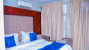 Een hotelkamer met een bed, twee stoelen, een raam met gordijnen en een airconditioning.