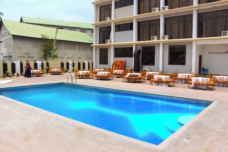 A swimming pool with lounge chairs and tables, surrounded by a building with multiple floors and air conditioning units.