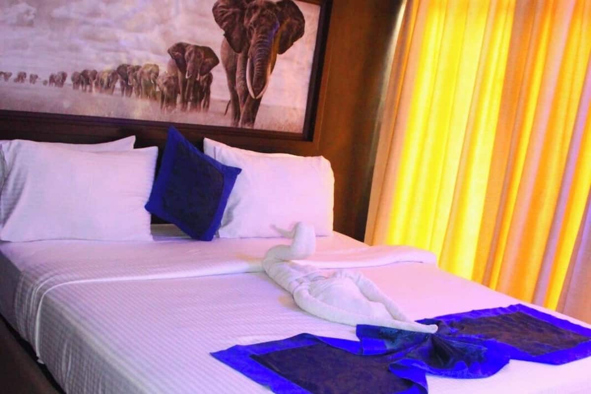 A hotel room with a large bed, white linens, a framed elephant painting, and a window with yellow curtains.