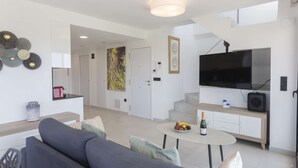 A modern living room with a grey sofa, a small round table with a bottle and fruit, a flat-screen TV, and a staircase leading to an upper level.