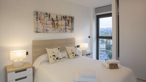 A neatly made bed with white linens and floral pillows, a bedside lamp, and a view of buildings through a large window.