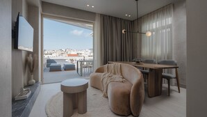 Un salon moderne avec un canapé, une table basse ronde et un coin repas avec des chaises. Un téléviseur est fixé au mur, et de grandes fenêtres offrent une vue sur la ville.