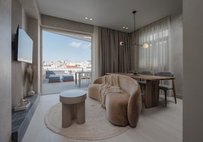 Un salon moderne avec un canapé, une table basse ronde et un coin repas avec des chaises. Un téléviseur est fixé au mur, et de grandes fenêtres offrent une vue sur la ville.