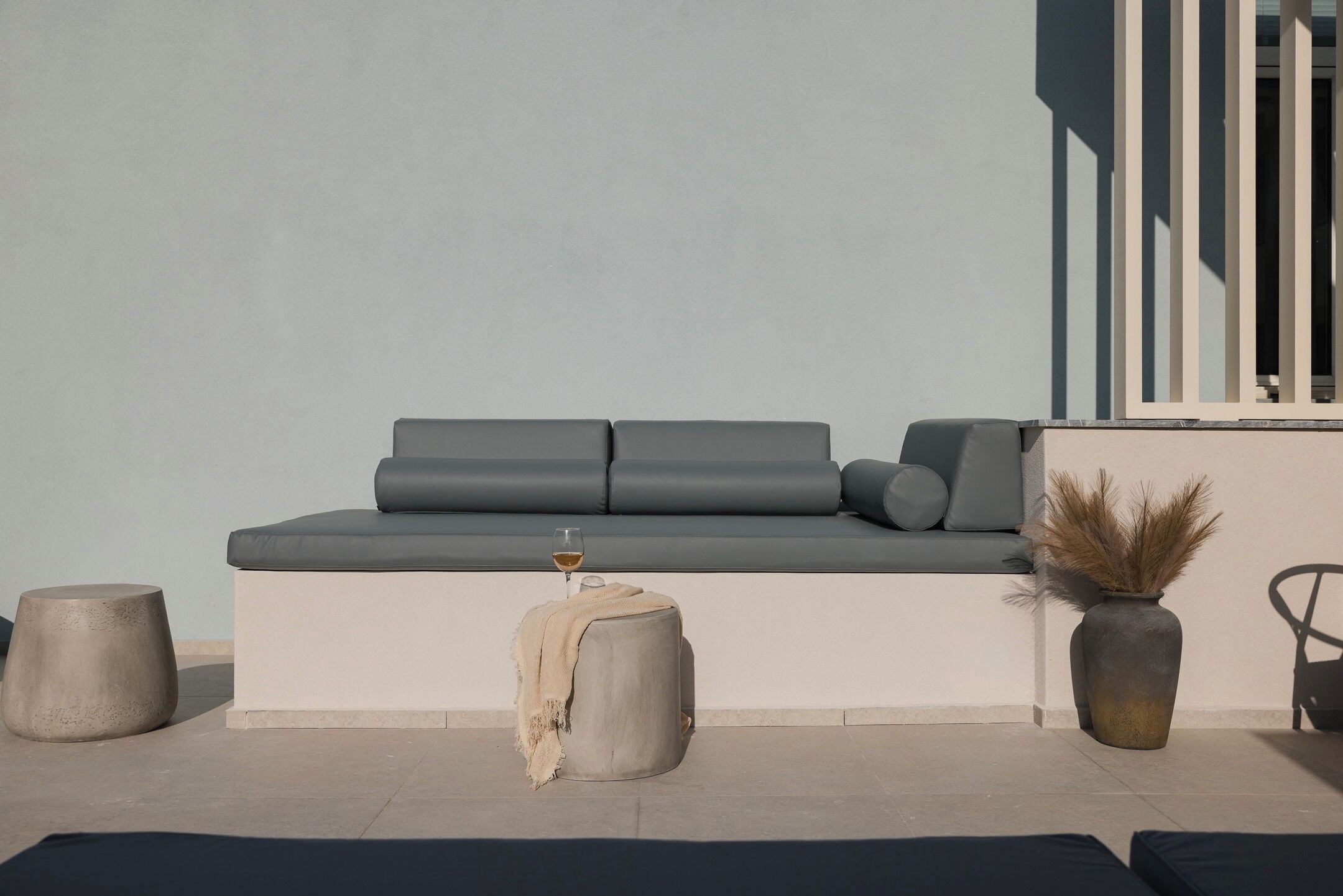 A modern outdoor seating area with a sofa, a round stool, and a vase with dried plants.