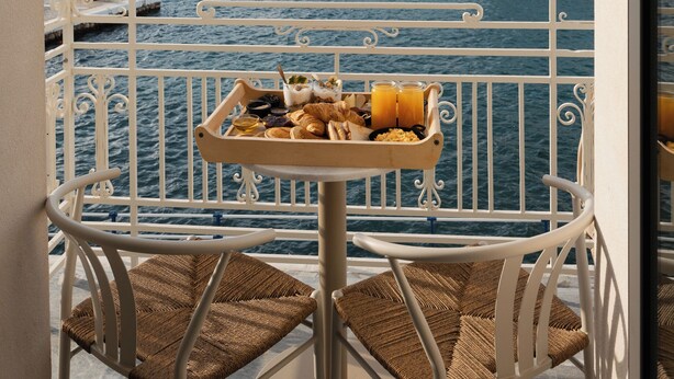 Un balcon avec des fauteuils en osier et un plateau de mets pour le petit-déjeuner, donnant sur un port avec des bâtiments en arrière-plan.
