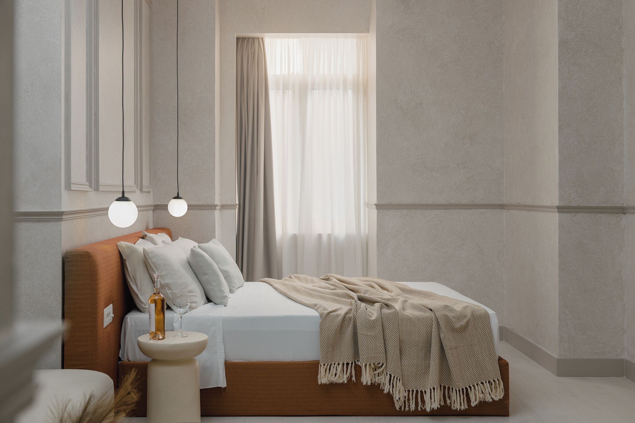 A modern bedroom with a wooden headboard, a bed with white linens, a beige throw blanket, and a bedside table with a bottle and glass.