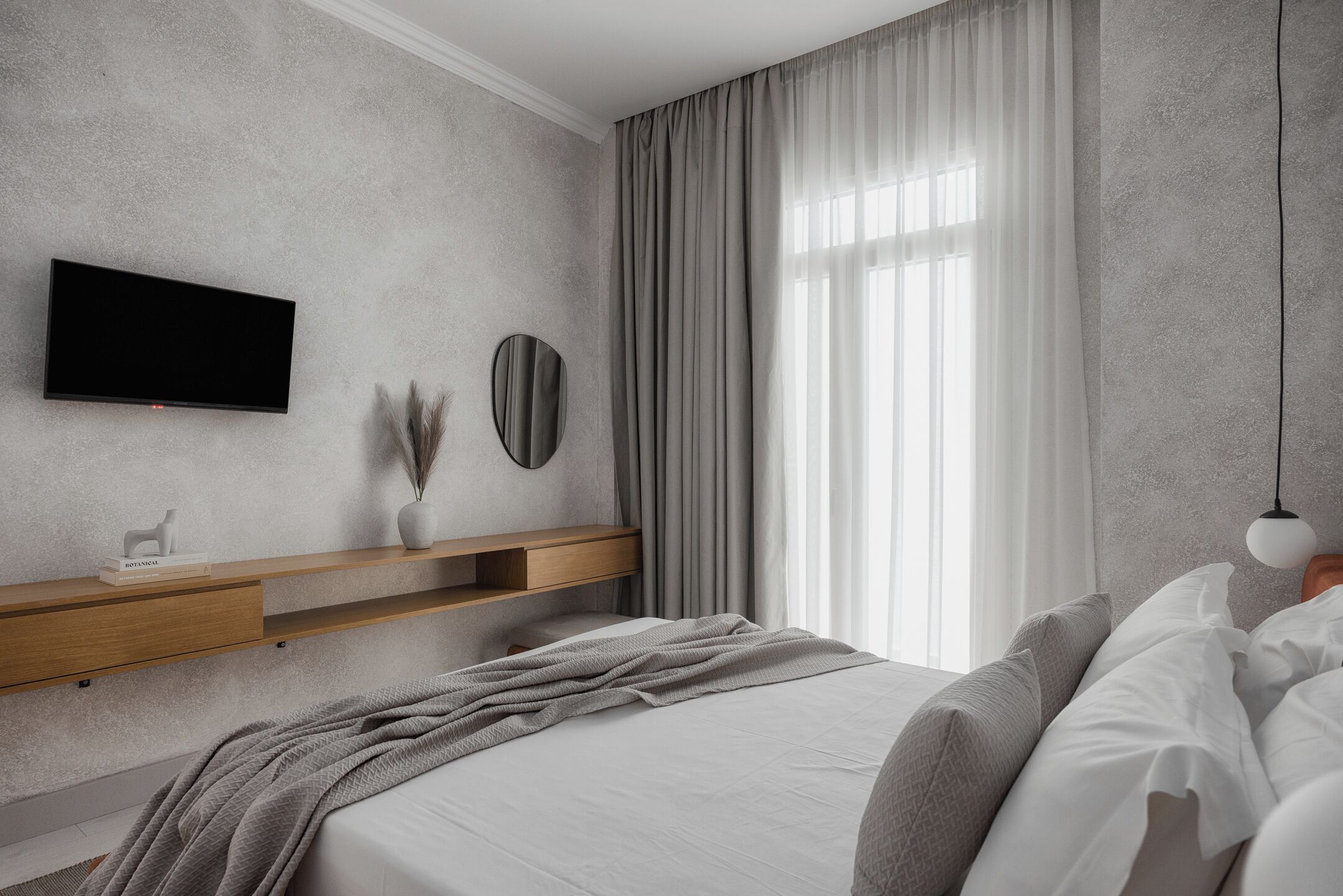 A modern bedroom with a flat-screen TV, a wooden shelf, a round mirror, and a large window with curtains.