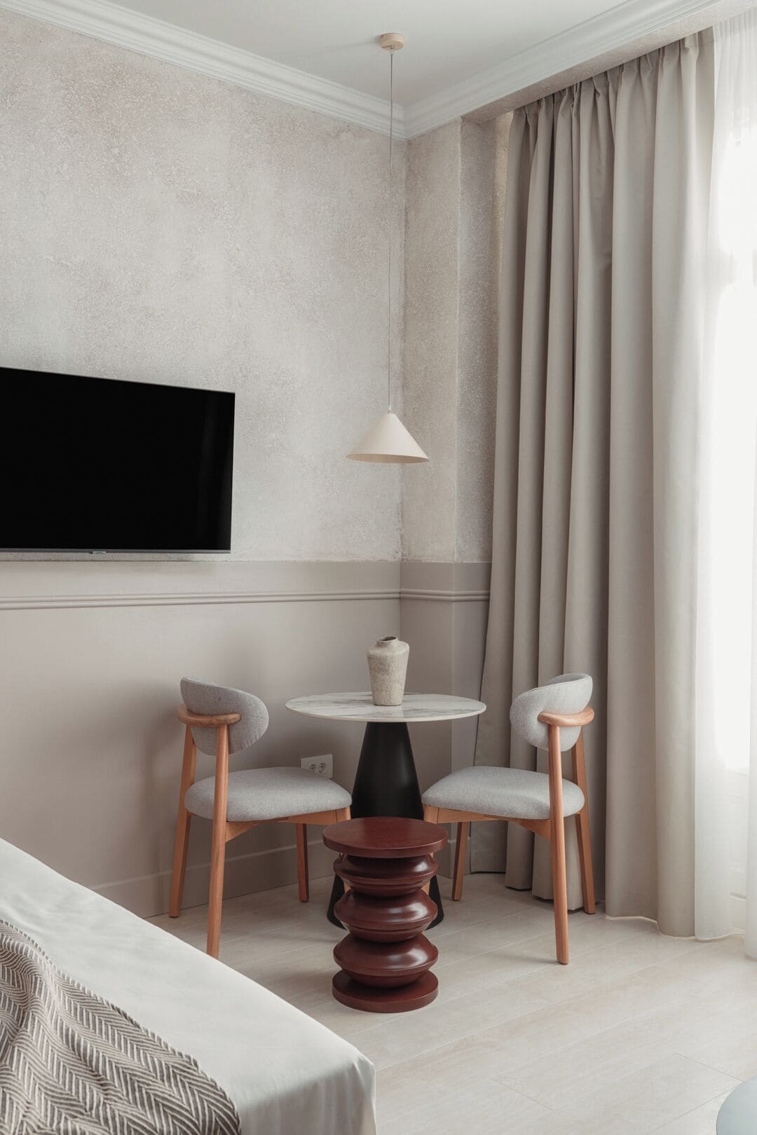 Moderni hotellihuone, jossa on suuri, seinälle asennettu litteänäyttöinen televisio, pyöreä pöytä kahdella tuolilla, ainutlaatuinen puinen sohvapöytä ja suuri ikkuna, jossa on verhot.