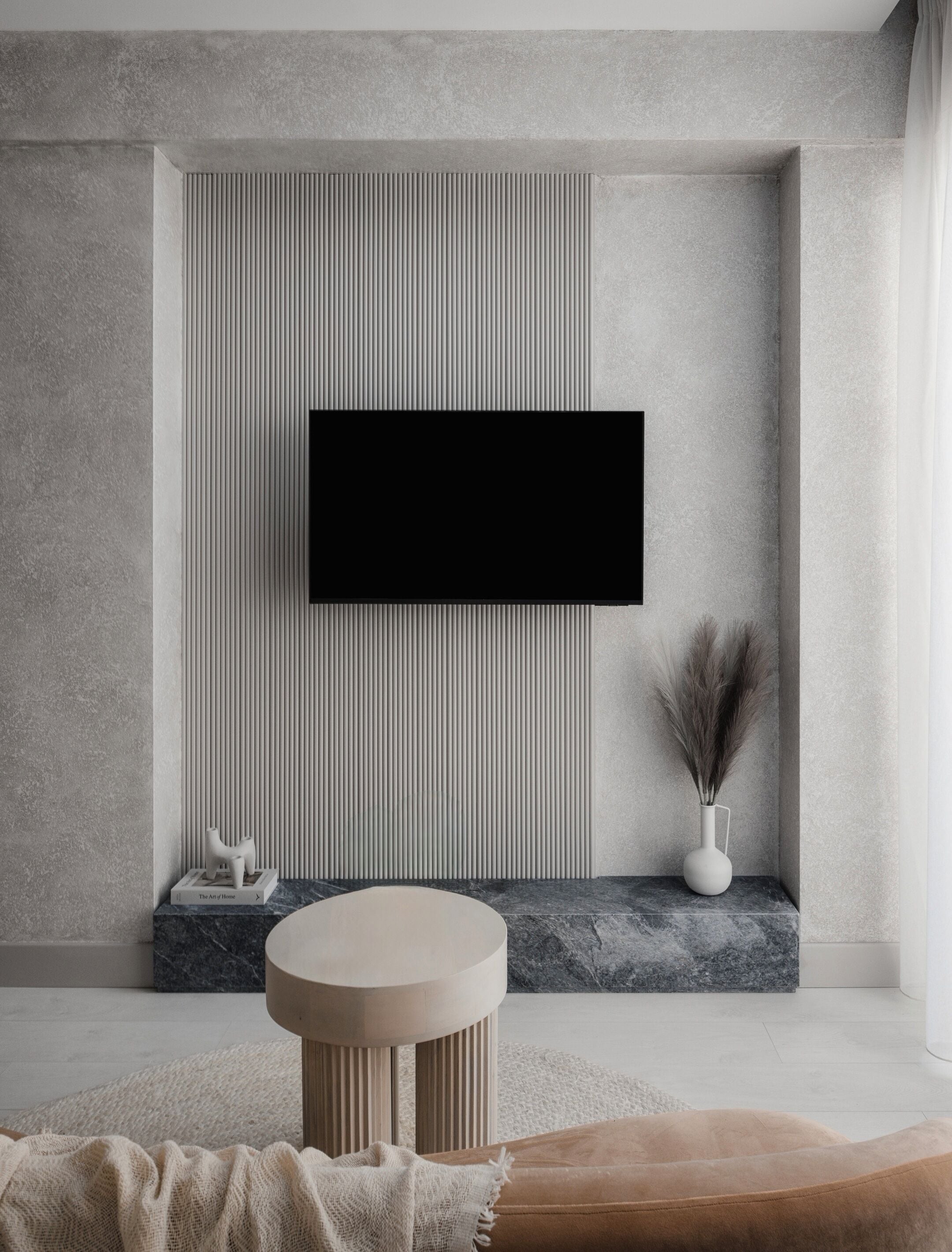 A modern living room with a flat-screen TV mounted on a textured wall, a round side table, a decorative vase, and a small sculpture.