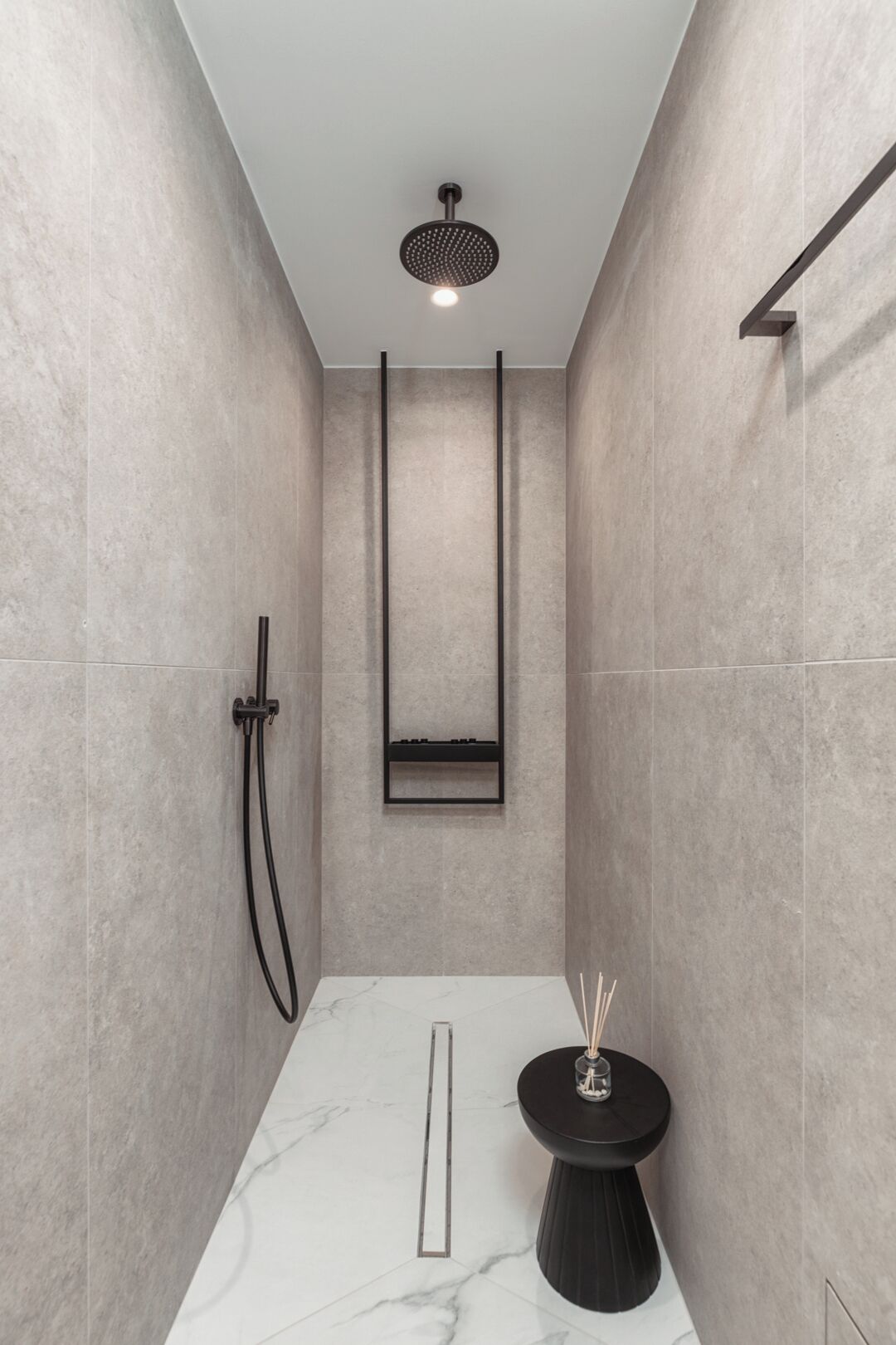 Un área de ducha moderna con un cabezal de ducha manual, un estante en la pared y una pequeña mesa con una vela.