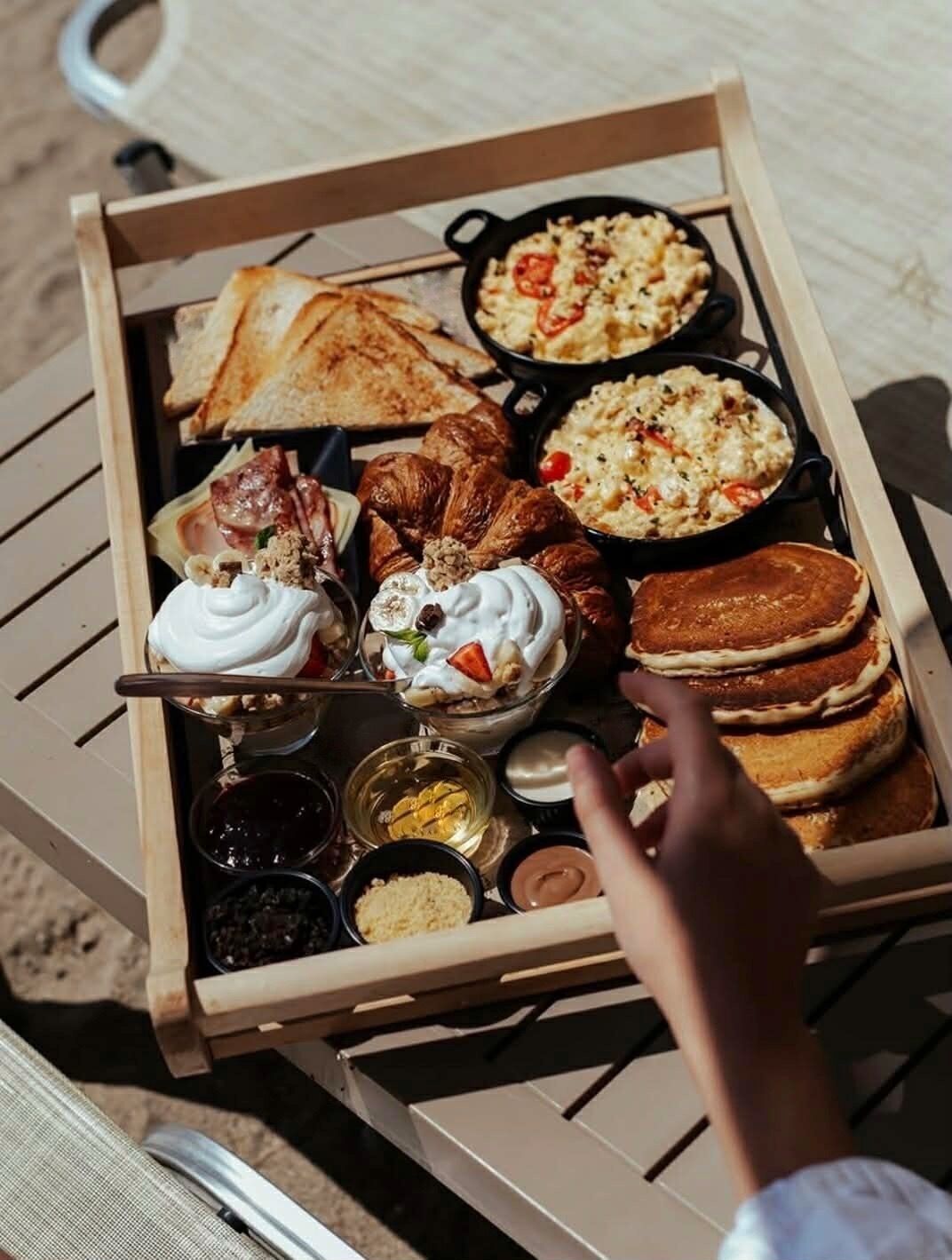 Un plateau en bois contenant divers éléments pour le petit-déjeuner, notamment des toasts, des œufs brouillés, des crêpes et un croissant.