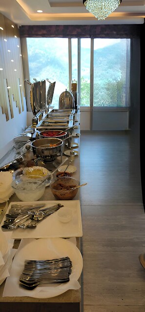 A buffet setup with various food items and utensils on a long table, with a view of greenery outside through large windows.