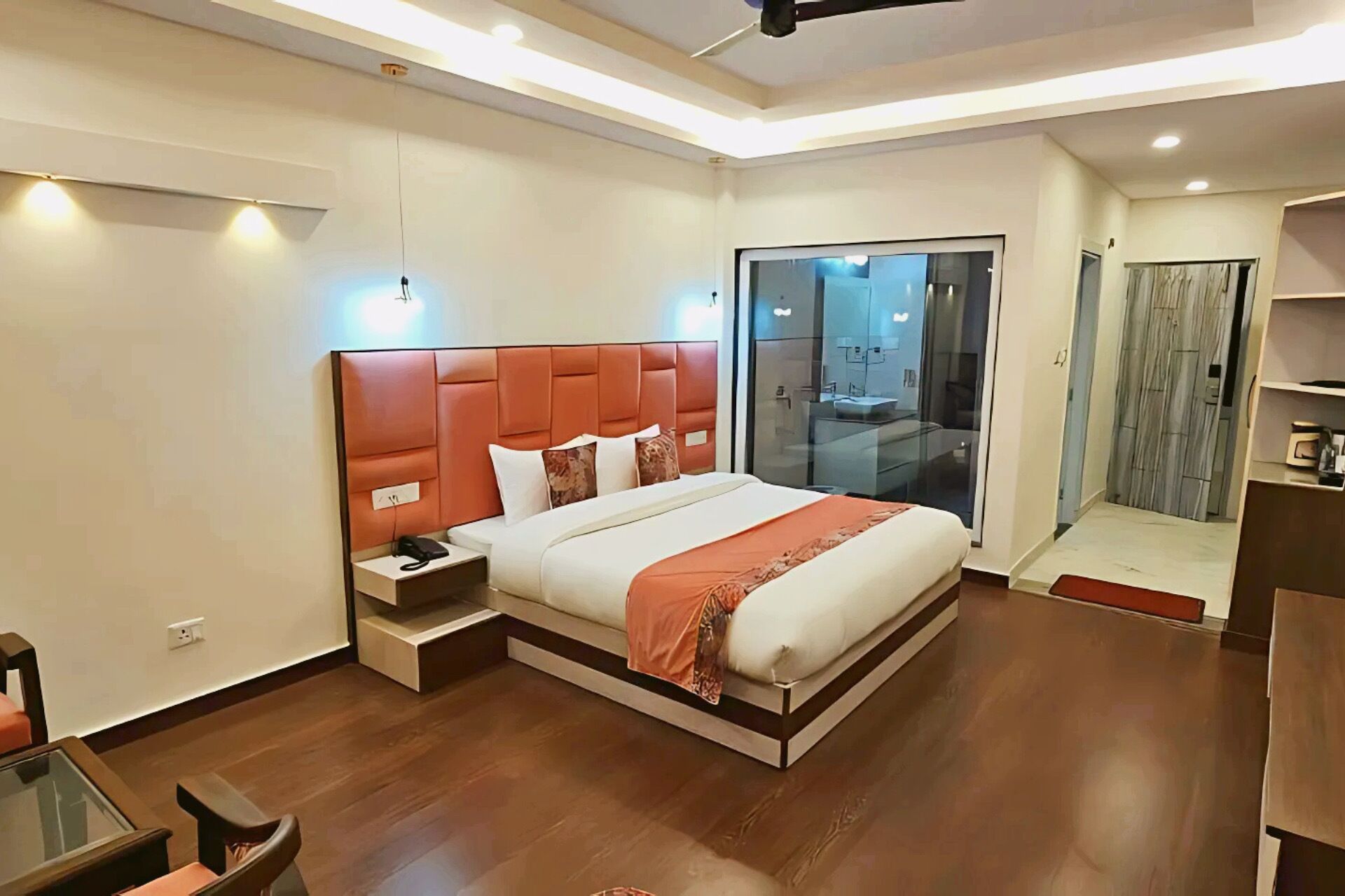 A modern hotel room with a large bed, a bedside table, a glass-enclosed bathroom, and a wooden floor.