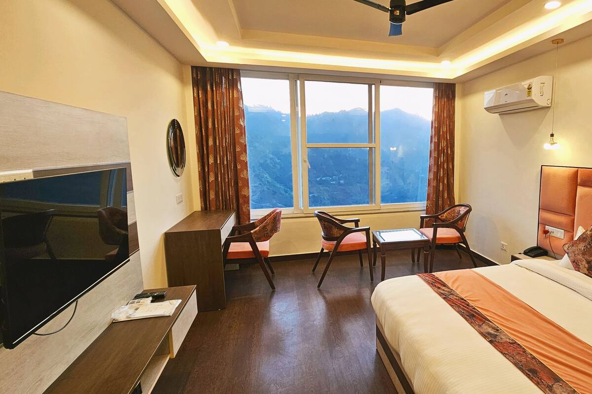 A hotel room with a bed, a desk with a TV, a chair, a table, and a window with a mountain view.