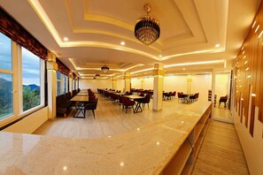 A spacious dining area with multiple tables and chairs, large windows offering a view of the outdoors, and a modern chandelier hanging from the ceiling.