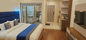 A hotel room with a large bed, a TV, a wooden shelf, and a bathroom visible through a glass door.