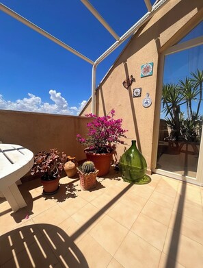 Eine sonnendurchflutete Terrasse mit Tisch, verschiedenen Blumentöpfen und einer Wand mit dekorativen Tellern.