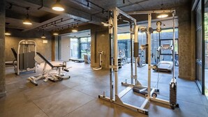 Un gimnasio con varios equipos de ejercicio, ventanas amplias y techo de hormigón.