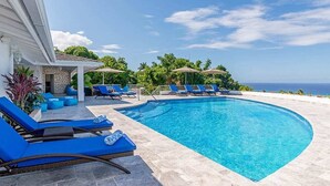 Una piscina con sillas reclinables azules y sombrillas, rodeada de follaje y vista al océano.