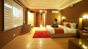 A hotel room with a large screen displaying a menu, a bed with a red rug, and a person sitting on the bed. - Udit Regency by west western kasol ( Central Heated and Air Cooled ) (Bhuntar)