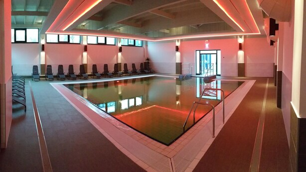 A modern indoor swimming pool with a glass barrier, lounge chairs, and a red-lit ceiling.