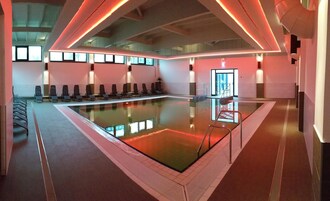 Una piscina interior moderna con barrera de vidrio, sillones de descanso y techo iluminado en rojo.