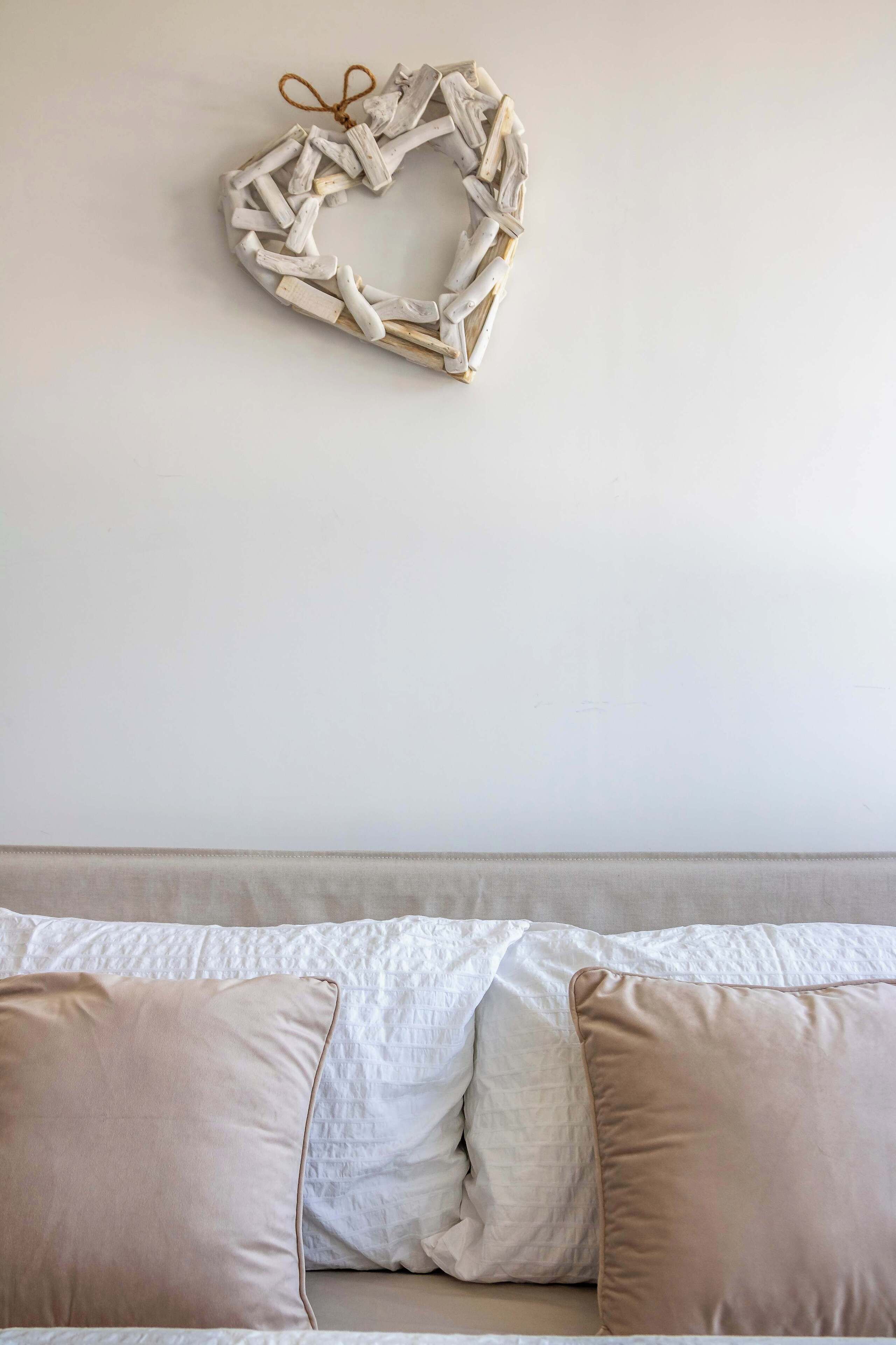 Une décoration en forme de cœur, en bois, fixée au mur au-dessus d’un lit agrémenté d’oreillers blancs et beige.