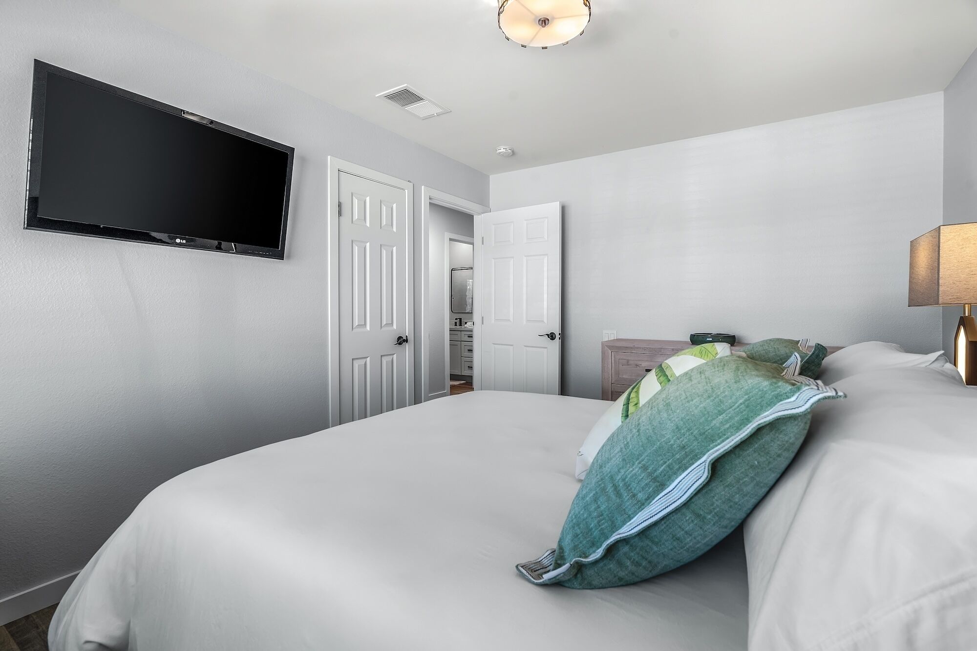 Une chambre d’hôtel dotée d’un téléviseur à écran plat fixé au mur, d’un lit avec une literie blanche et un oreiller vert, d’une lampe de chevet et d’une porte donnant accès à une salle de bain.