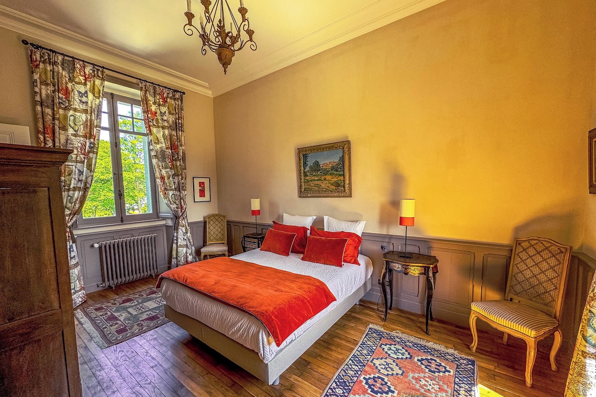 Een slaapkamer met een groot bed, een stoel, een tafel, een kroonluchter en een raam met gordijnen.