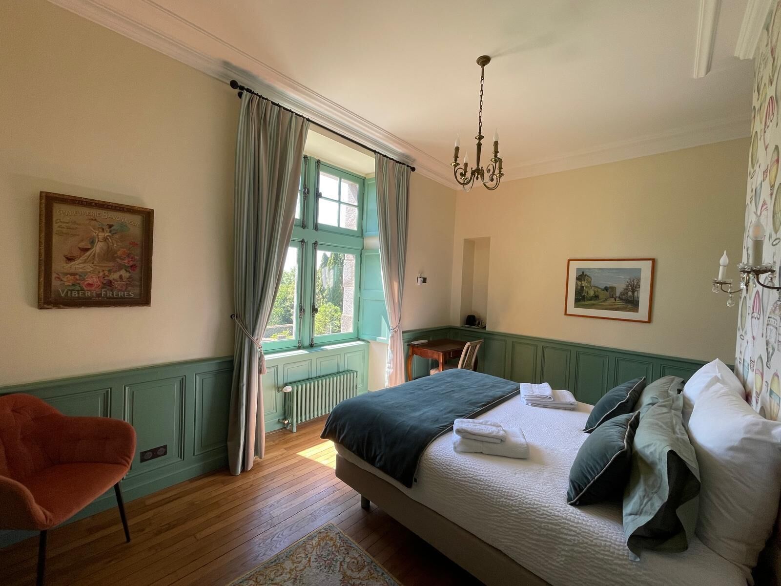 A bedroom with a large bed, a chair, a radiator, a window with curtains, and a chandelier.