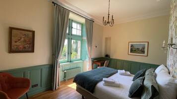 A bedroom with a large bed, a chair, a radiator, a window with curtains, and a chandelier.