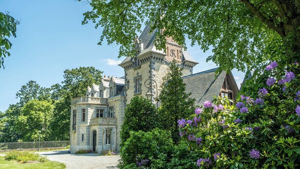 Een historisch gebouw van steen met een toren en een kleinere vleugel, omringd door weelderig groen en paarse bloemen.