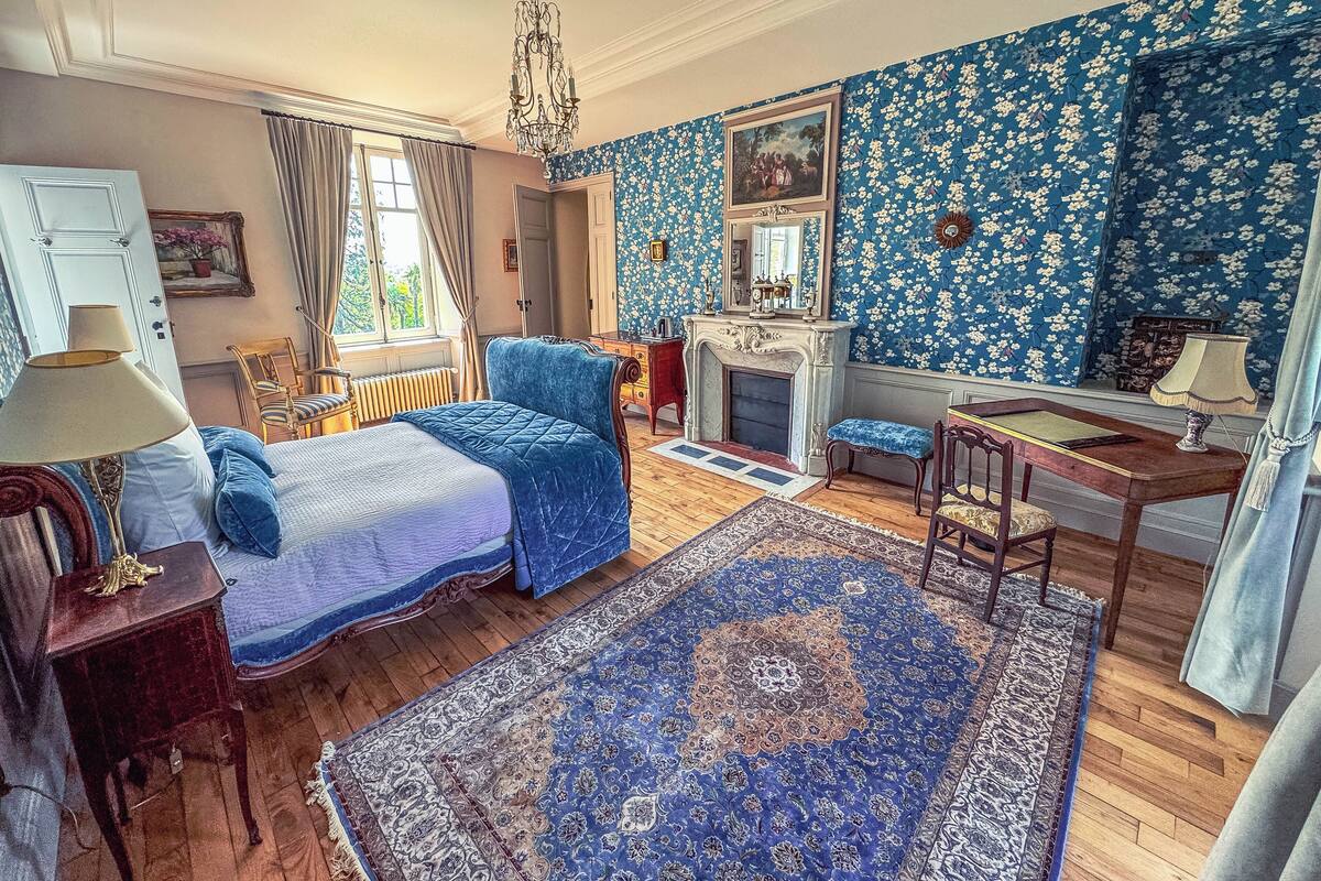 Een klassieke slaapkamer met een bed, een open haard, een bureau en een groot raam.