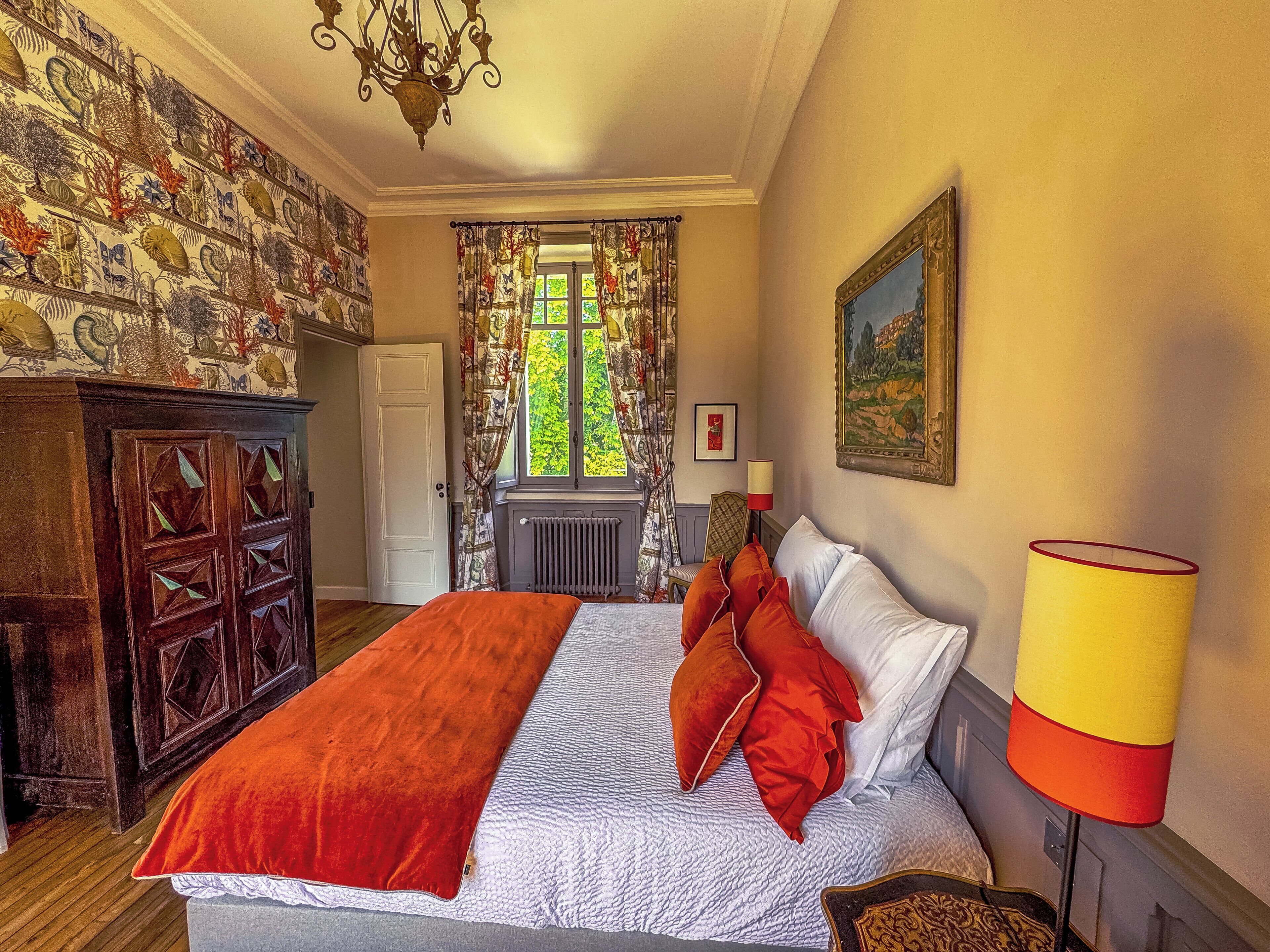 A bedroom with a large bed, a wooden wardrobe, a radiator, a window with curtains, and a chandelier.