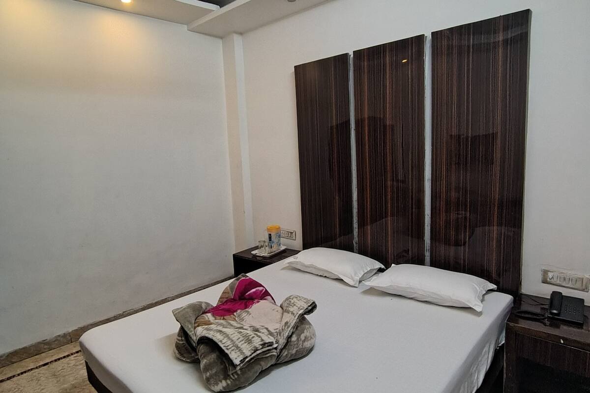 A bedroom with a bed, a bedside table, a nightstand, a wall-mounted light fixture, and a large window with curtains.