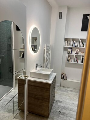 A modern bathroom with a glass shower enclosure, a white vanity with a marble countertop, a round mirror with integrated lighting, and a built-in bookshelf with books. - Oceanfront Getaway – Terrace, View & Free Parking (Ajaccio)