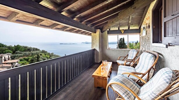 A wooden balcony with wicker furniture and a wooden table, overlooking a scenic view of the sea and hills. - Antico Fienile Belvedere (Gardone Riviera)