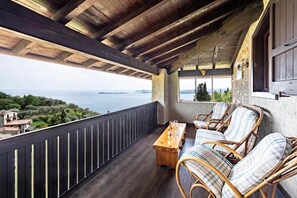 A wooden balcony with wicker furniture and a wooden table, overlooking a scenic view of the sea and hills. - Antico Fienile Belvedere (Gardone Riviera)