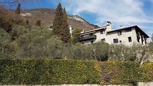 A stone building with a balcony, surrounded by dense shrubbery and trees, with a mountain in the background. - Antico Fienile Belvedere (Gardone Riviera)