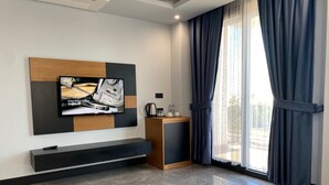 Una habitación de hotel moderna con un televisor de pantalla plana grande montado en una pared de madera y panel negro, un mueble de madera pequeño, una cafetera y un ventanal con cortinas oscuras.