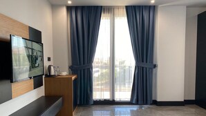 Una habitación de hotel moderna con un televisor grande montado en la pared, una mesita consola de madera, una pequeña kitchenette y un amplio ventanal con cortinas azules.
