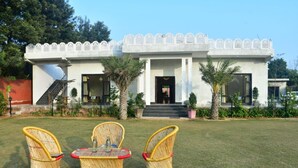A two-story building with a white facade, a balcony with intricate lattice work, and a front entrance with steps. There are three wicker chairs and a small table in the foreground. - Aroma Green Resort (Gurugram)
