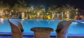 A poolside area with wicker chairs and tables, surrounded by palm trees and outdoor lighting. - Aroma Green Resort (Gurugram)