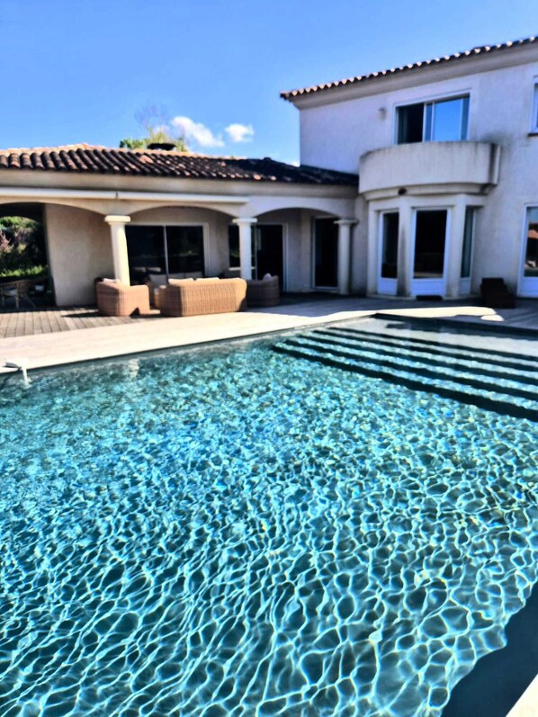Una piscina con agua cristalina, un área de patio con mobiliario y un edificio con techo de tejas y columnas.