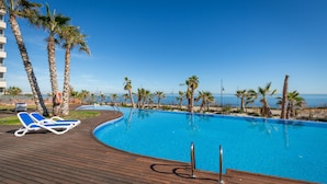 En klarblå pool med ett trädäck och solstolar, omgiven av palmer och med utsikt över havet.