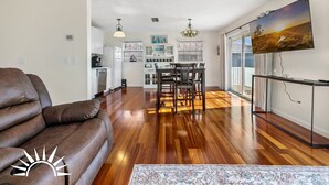 A spacious living room with a brown leather sofa, a dining area with a table and chairs, a kitchen with white cabinets, and a large window with blinds.